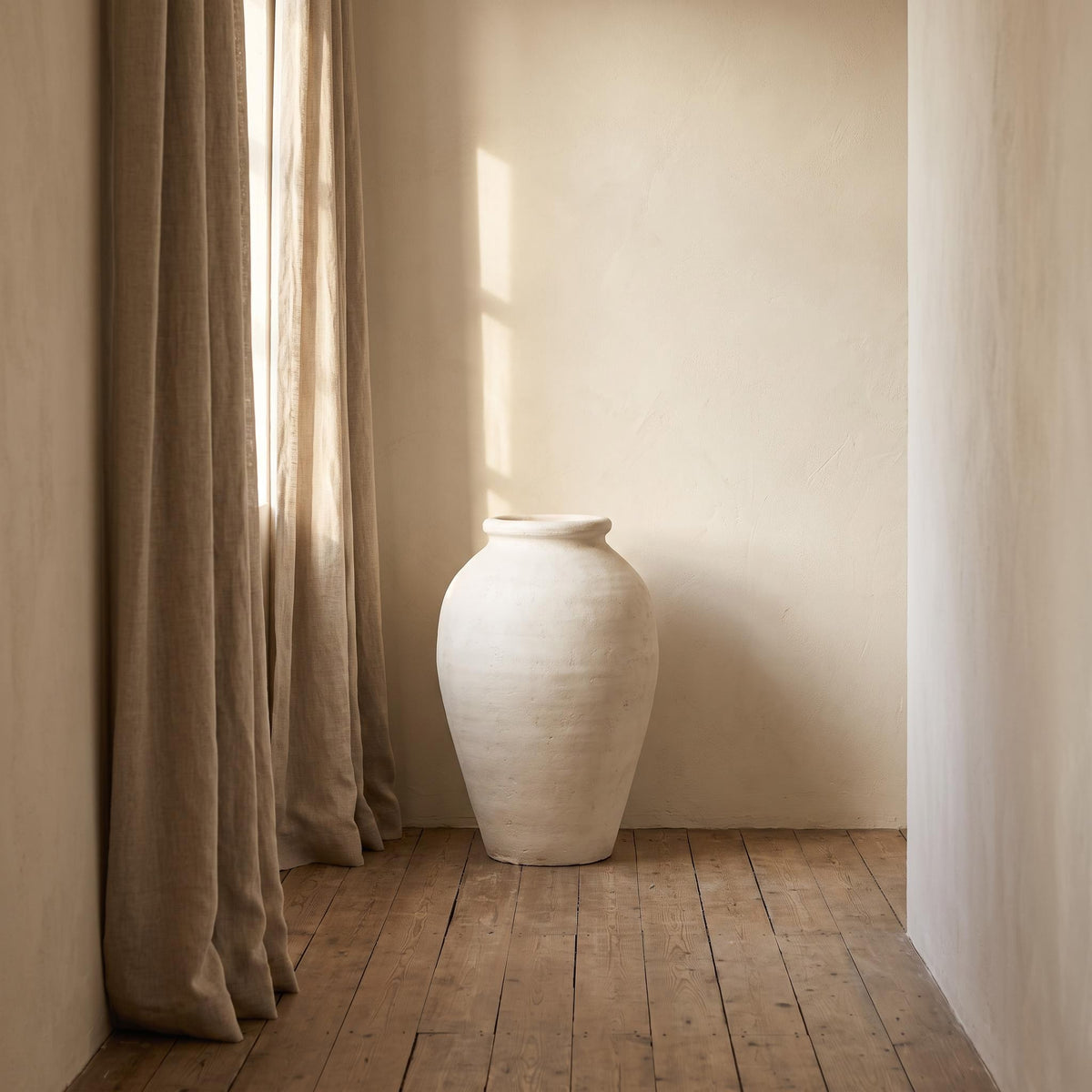 Bianca vase empty on a hallway floor beside a linen curtain in afternoon light
