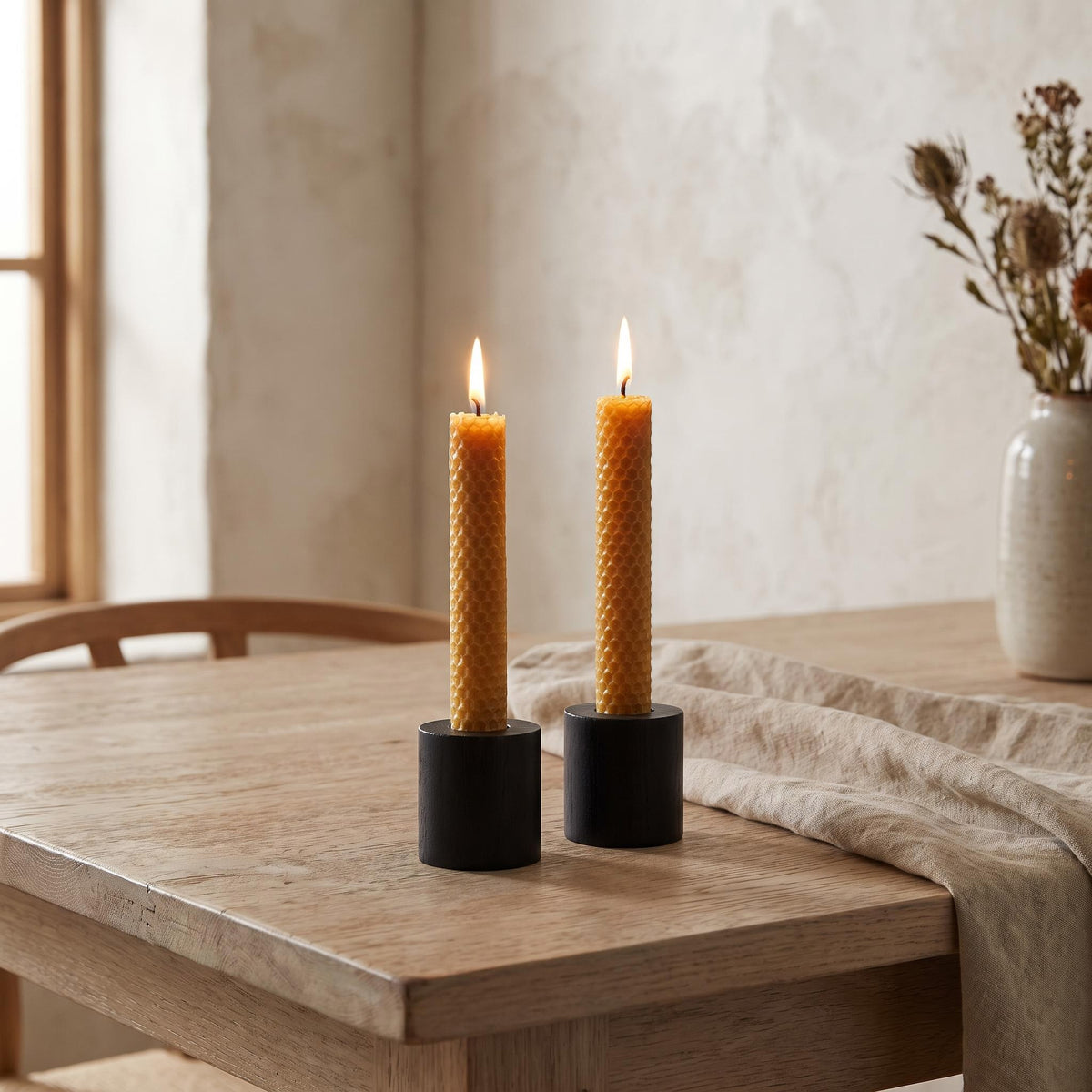 Panal beeswax candles lit in black wooden holders on a pale oak dining table